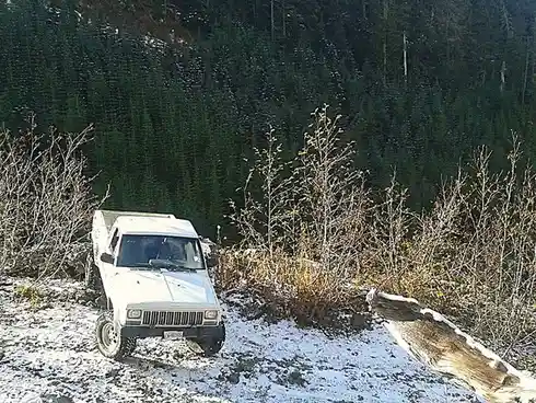A white pickup truck is parked on a snowy, forested hillside with sparse trees and brush. The vehicle appears to be…