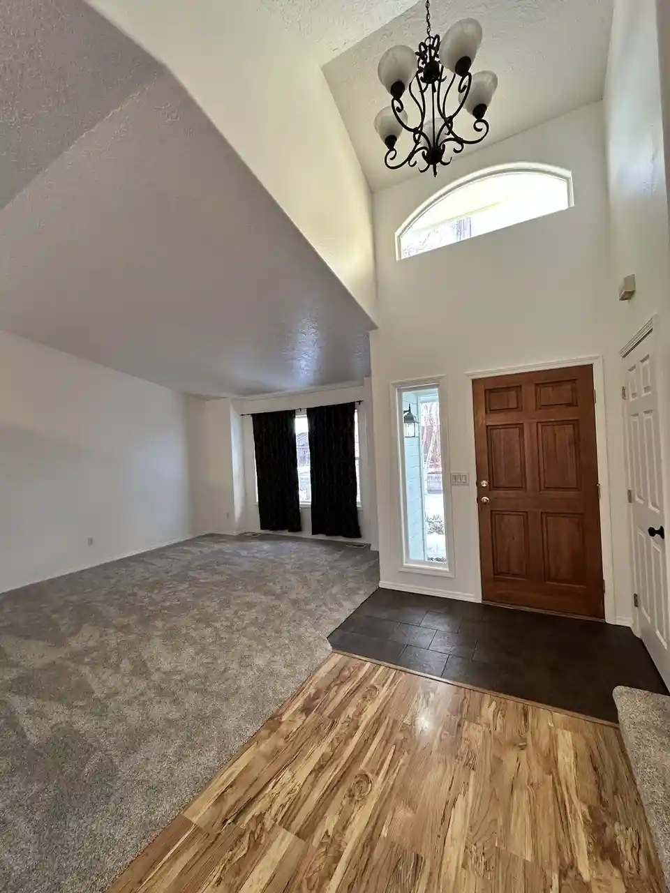 An empty, spacious room in a house features three distinct flooring types: light wood laminate in the foreground, dark gray…