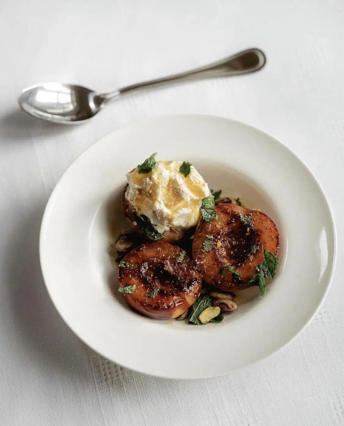A white bowl holds three caramelized peach halves topped with a scoop of vanilla ice cream and garnished with mint leaves…
