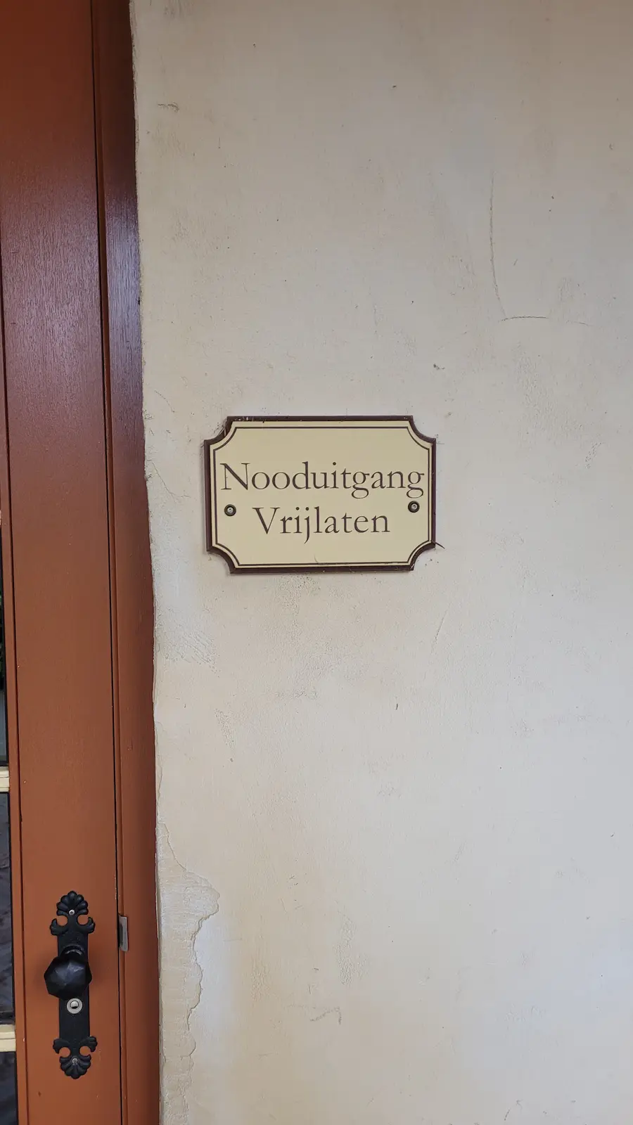 A close-up photo shows a cream-colored, framed sign mounted on a textured beige wall next to a brown wooden door with a…