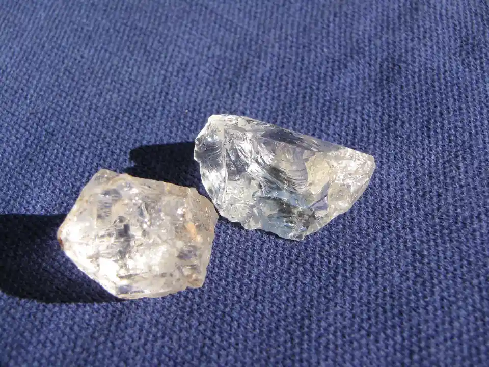 Two rough, uncut, clear gemstones resembling diamonds rest on a textured blue fabric surface. The overlaid text reads 'two…