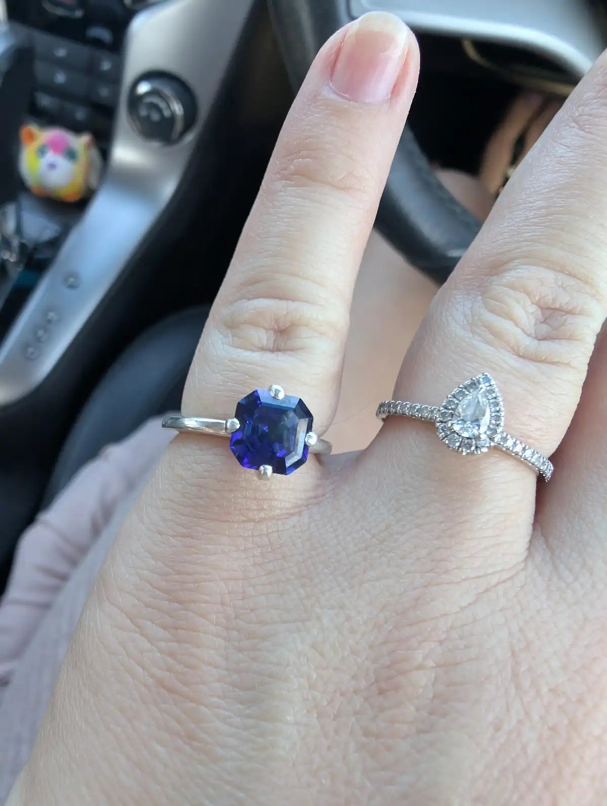 A close-up photo shows a person's hand inside a car, displaying two rings: one with a large, deep blue sapphire on the index…