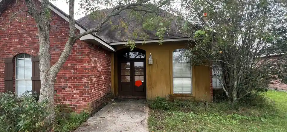 A photo of a modest brick and stucco house with a concrete walkway, surrounded by trees and overgrown grass. A red circle is…