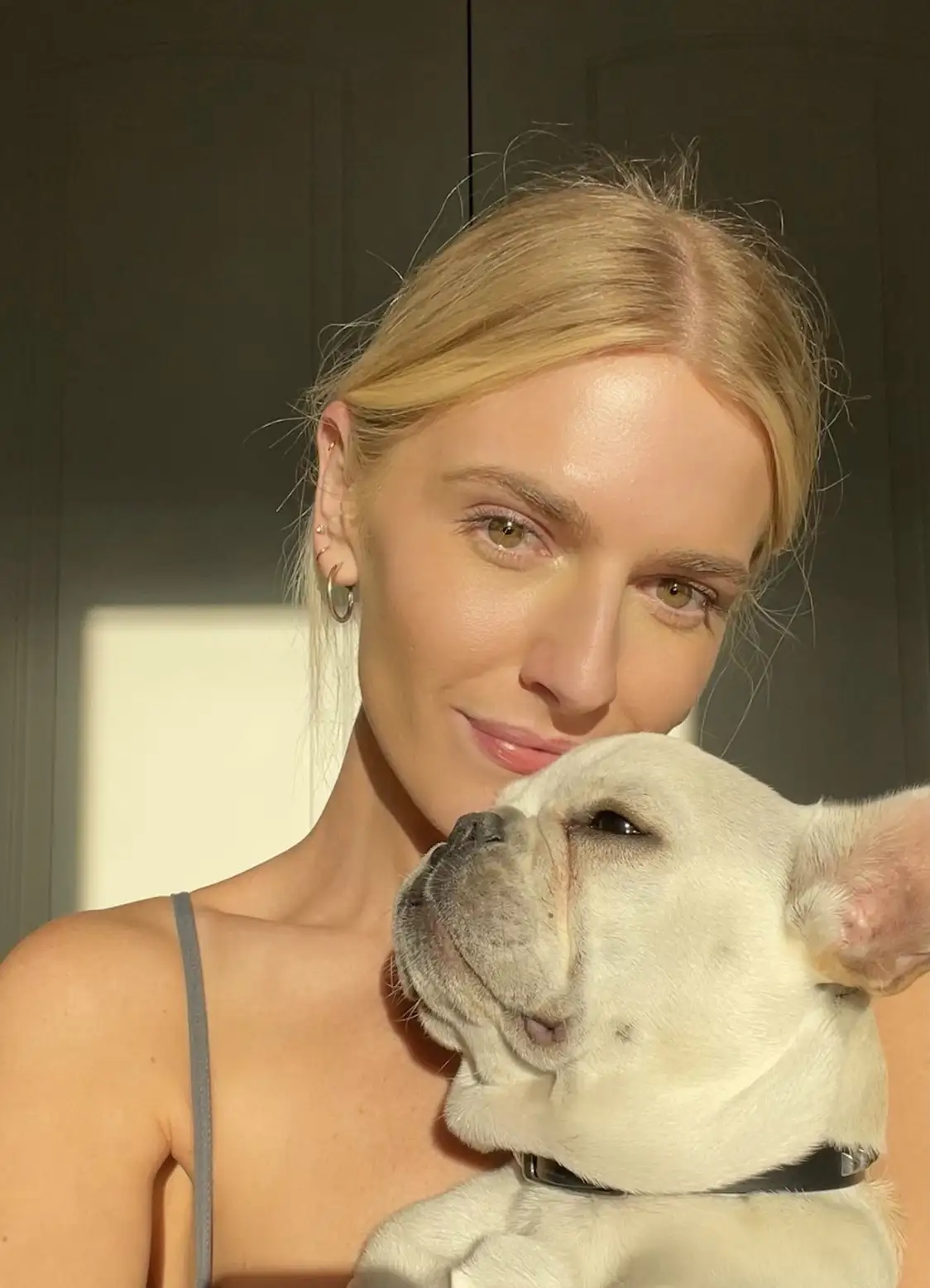 A woman with blonde hair and a gray tank top is holding a white French bulldog close to her chest. The dog is looking up at…