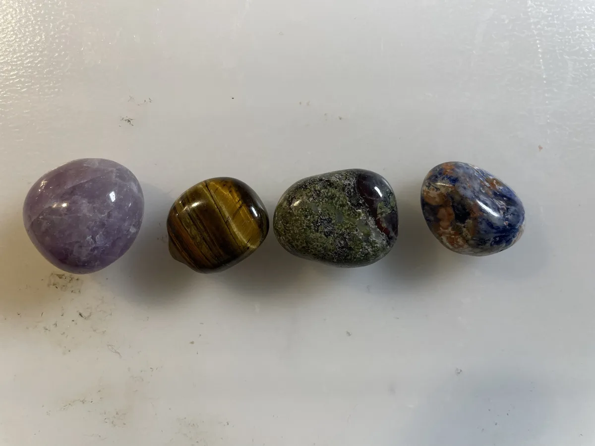 Four polished, colorful stones—purple amethyst, striped tiger eye, greenish speckled stone, and blue/orange patterned…