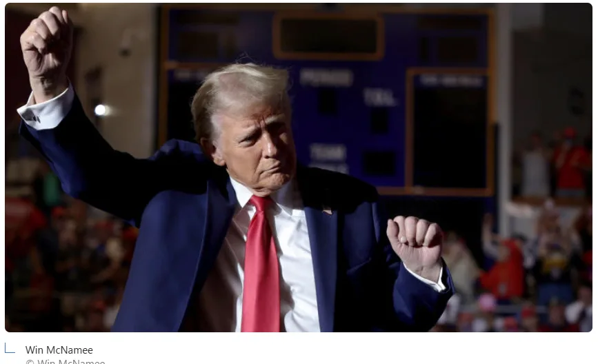 a photo of Donald Trump in a dark suit and red tie, raising one fist while his other hand is clenched, with a crowd and…