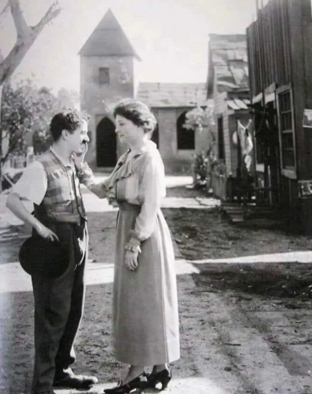 A black-and-white vintage photo shows a woman in a long dress gently touching the cheek of a man in a vest and bowler hat…