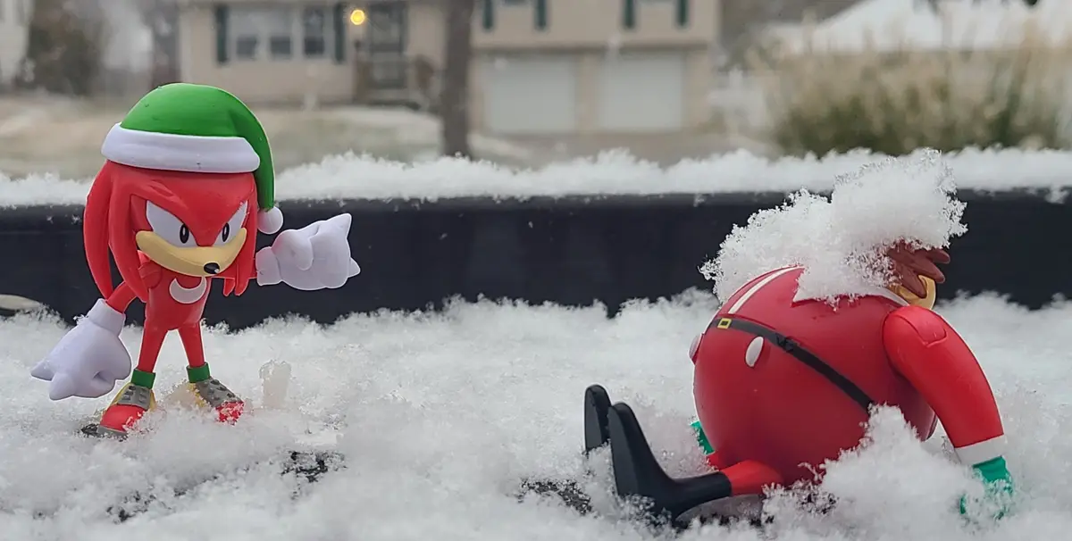 Two Sonic the Hedgehog action figures are in a snowy outdoor scene. One figure, Knuckles, stands upright wearing a green and…
