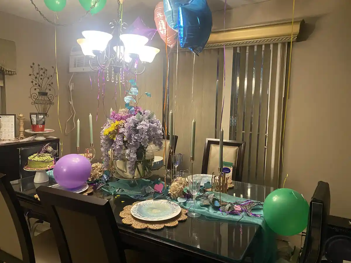 A dining room is elaborately decorated for Mardi Gras with colorful balloons, streamers, a festive tablecloth, candles, and…