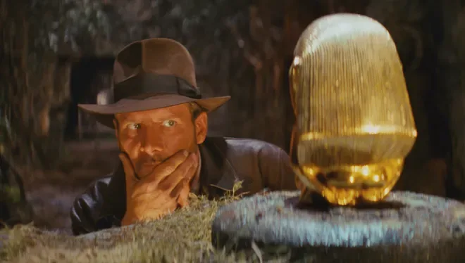 Indiana Jones, wearing his iconic fedora and leather jacket, peers cautiously over a grassy ledge at a large, ornate golden…
