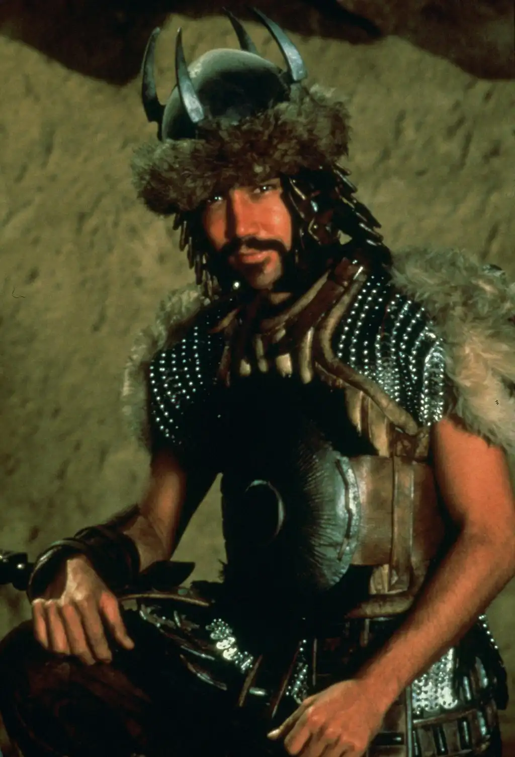A man in elaborate, spiked, fur-lined armor and helmet sits with a confident, slightly smug expression, overlaid with the…