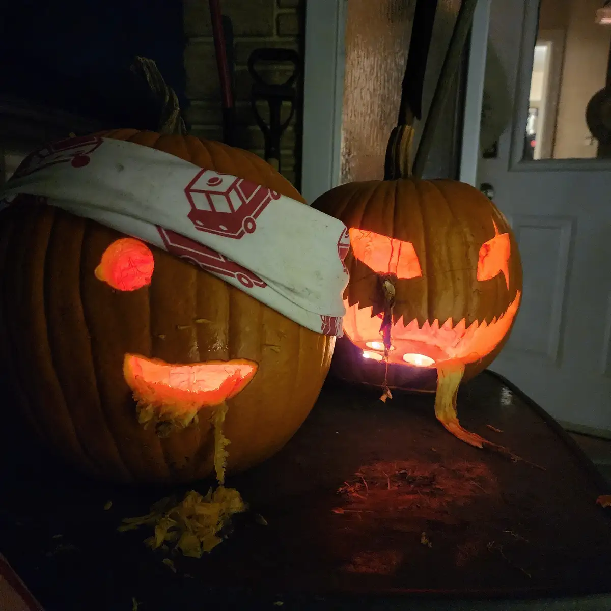Two carved Halloween pumpkins sit on a porch at night, illuminated from within. The pumpkin on the left wears a white cloth…