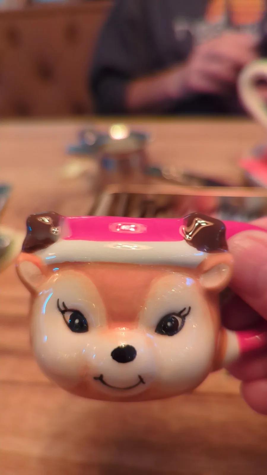 A close-up photo shows a hand holding a ceramic mug shaped like a cartoon fox with a pink hat and a smiling face. The…