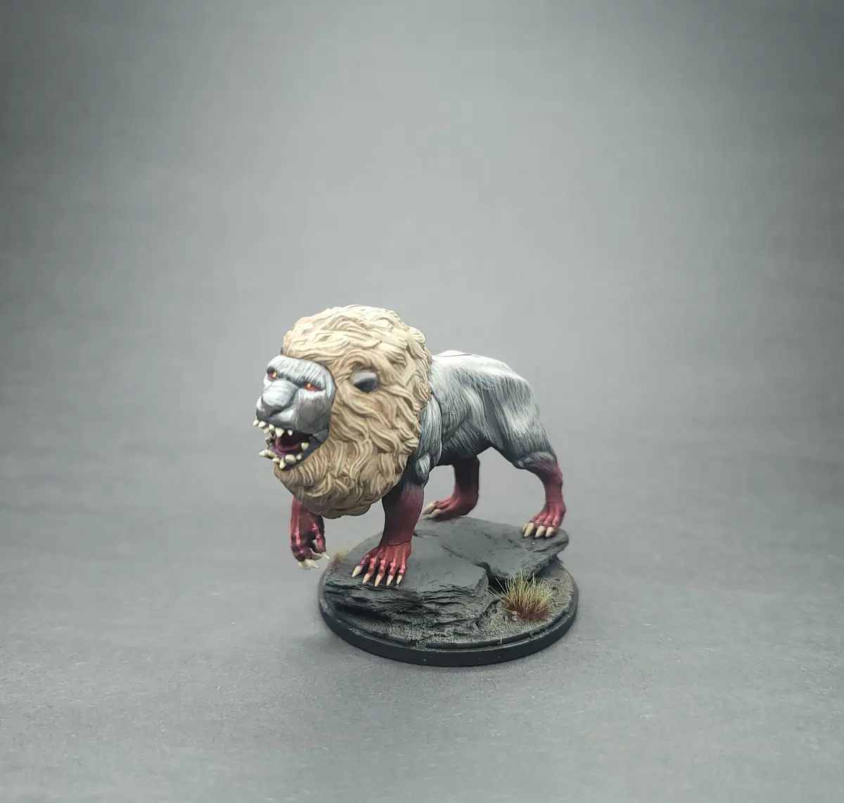 A painted miniature of a lion with grey fur, a tan mane, and red paws stands on a rocky base against a grey background. The…