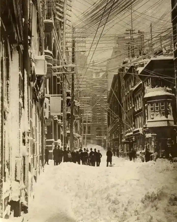 A sepia-toned historical photograph shows a snow-covered city street with tall buildings on both sides, thick snow piled…