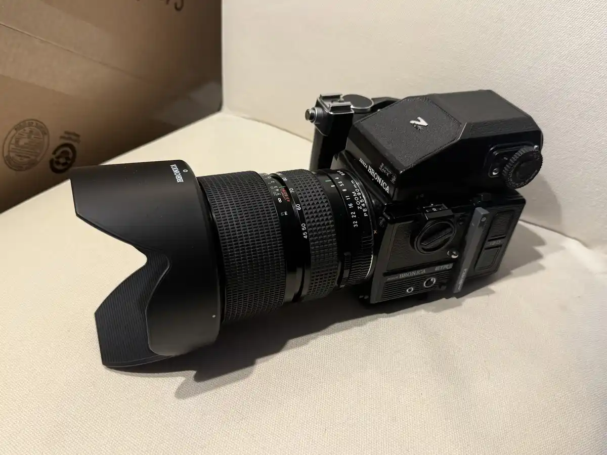 A black Zenit Bronica ETRSi medium format film camera with a large zoom lens and lens hood rests on a white textured…
