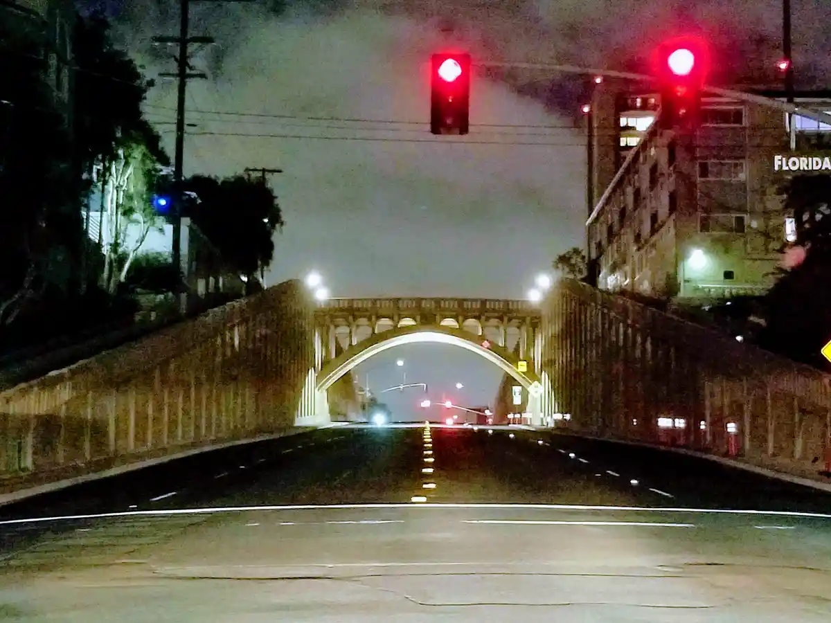 A nighttime dashcam view from a car stopped at a red traffic light, facing a large arched bridge ahead. Two red traffic…