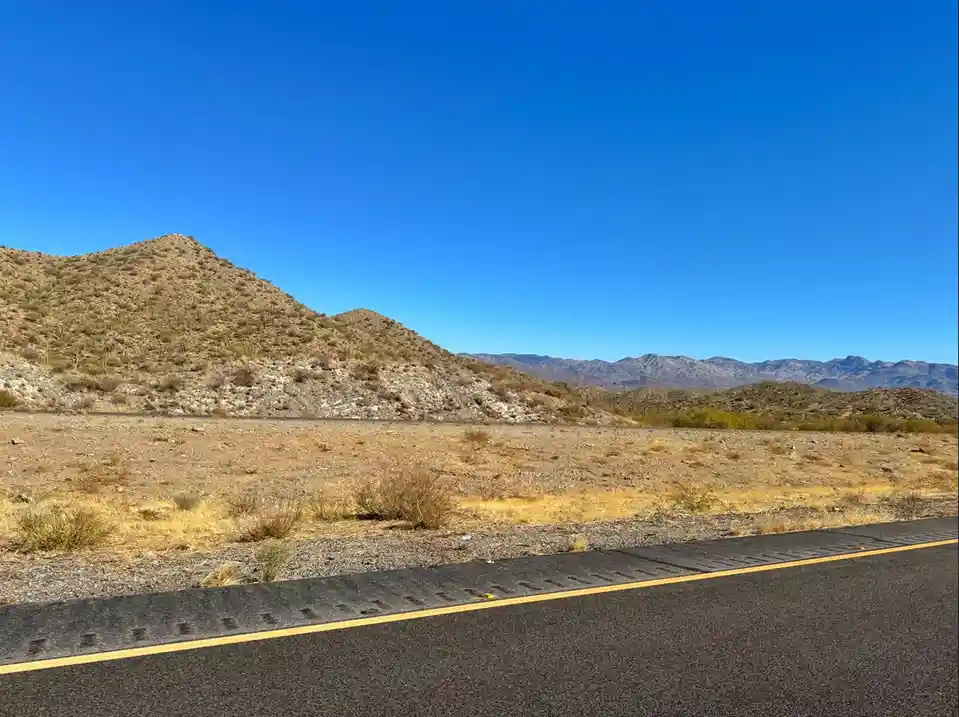 A wide desert landscape with a paved road in the foreground, dry brush, distant mountains, and a clear blue sky. Overlaid…