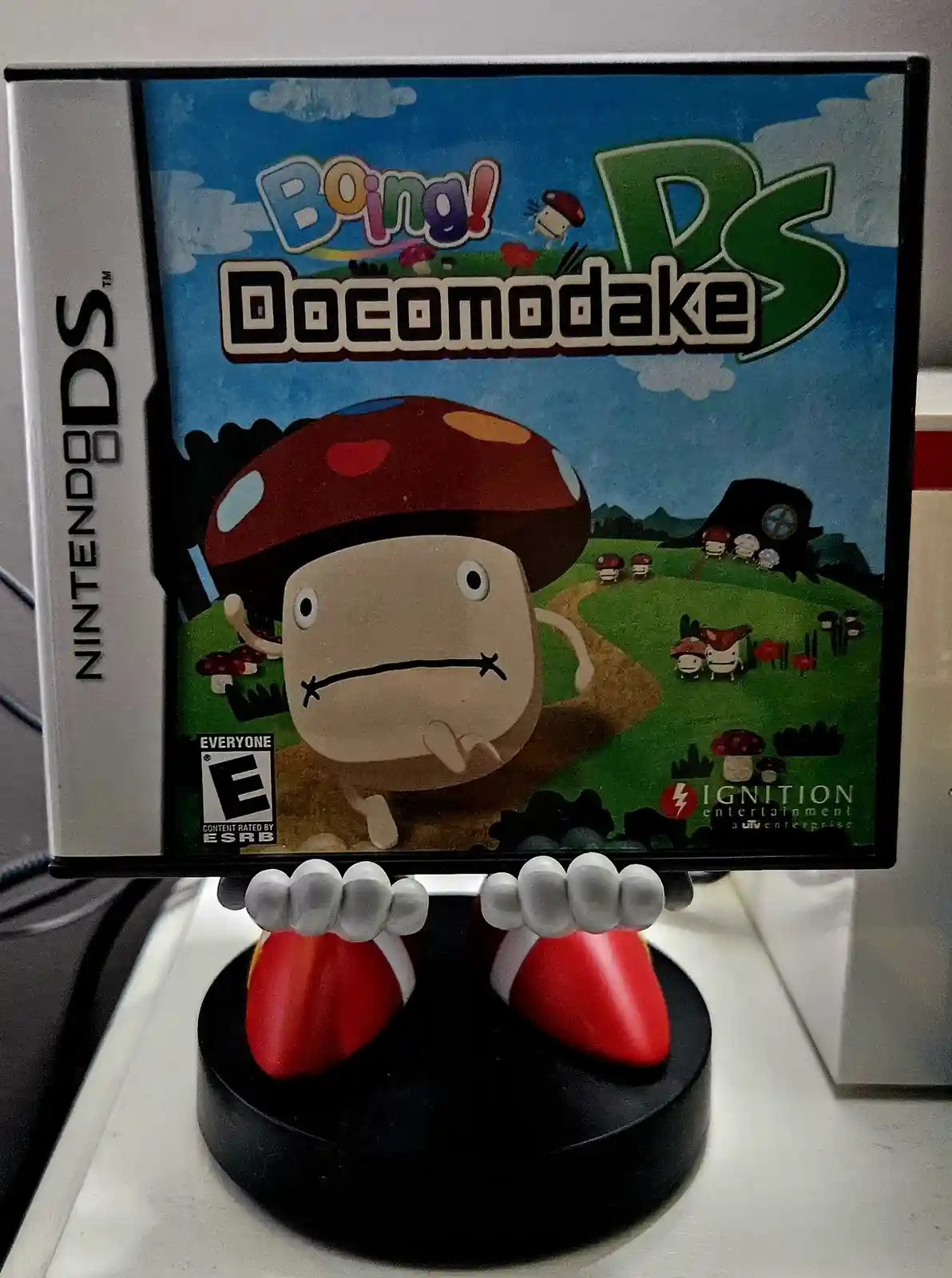 A Nintendo DS game case for 'Boing! Doconodake' is displayed on a stand shaped like Mario's feet, creating a humorous…