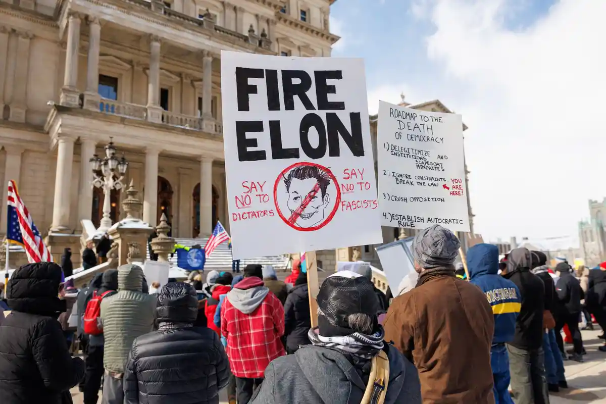 A protest scene with people gathered in front of a large classical building, holding signs. One prominent sign reads 'FIRE…