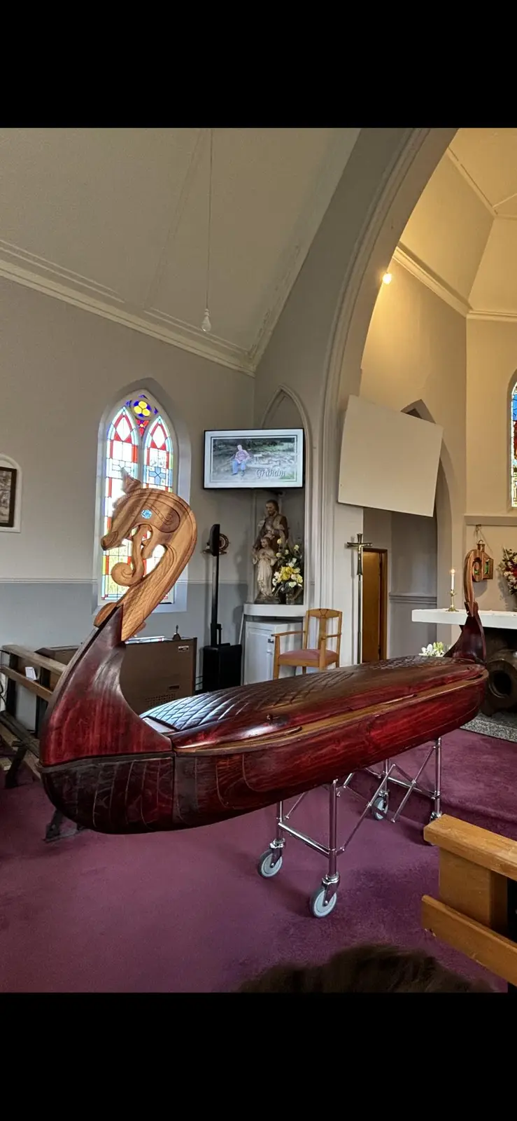 A polished wooden Viking-style funeral boat coffin with dragon-head carvings sits on wheeled metal stands inside a church…