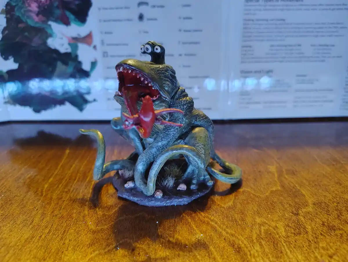 A small, green, multi-limbed monster figurine with a wide-open mouth and two large eyes on a stalk is shown on a wooden…