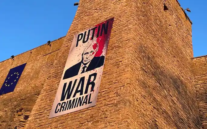 A large banner is affixed to a brick building, featuring a portrait of Vladimir Putin with red splatter effects, and the…
