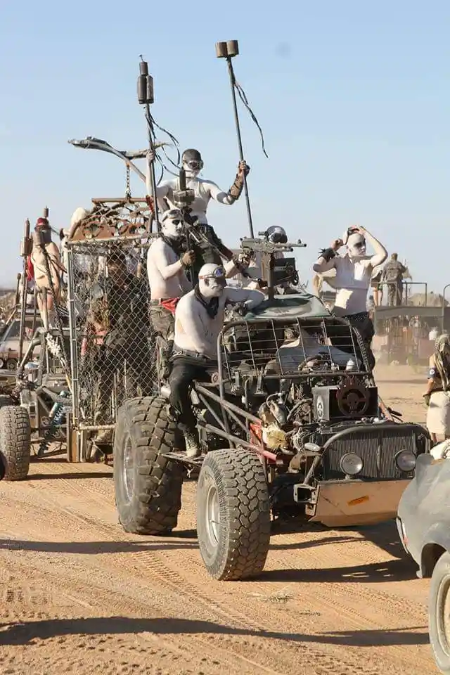 A group of people with white face paint and goggles ride atop a heavily modified, rugged, desert vehicle with large tires…