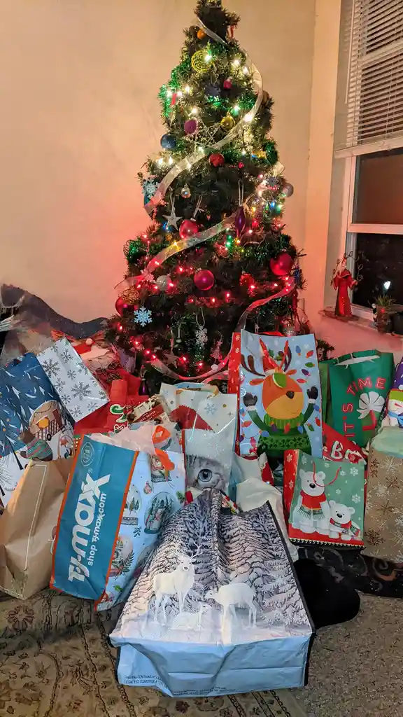 A brightly lit, decorated Christmas tree stands behind a large pile of holiday shopping bags, including recognizable ones…