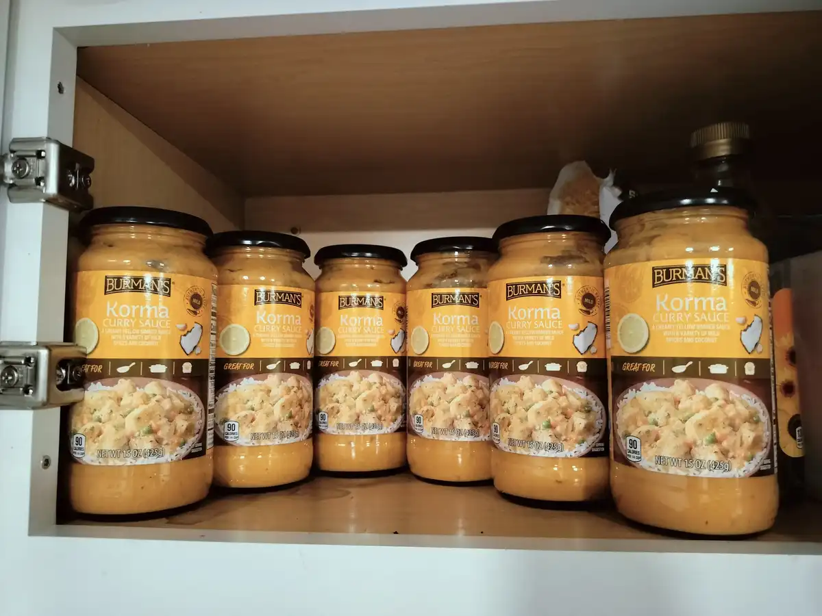 A kitchen cabinet shelf holds six identical jars of Burman's Korma Curry Sauce, arranged neatly in a row. The labels clearly…