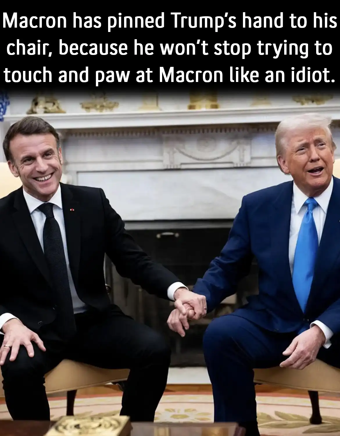Two men in suits sit in chairs facing each other, shaking hands. The man on the left, wearing a black suit, has his other…