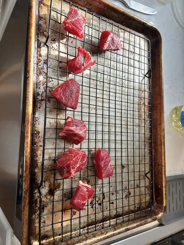 A close-up photo shows several raw, red meat chunks arranged on a black wire rack inside a well-used, browned metal baking…