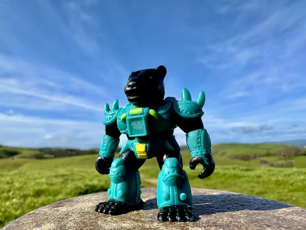 A black panther-like action figure with teal armor stands on a rock against a backdrop of green hills and blue sky. The…
