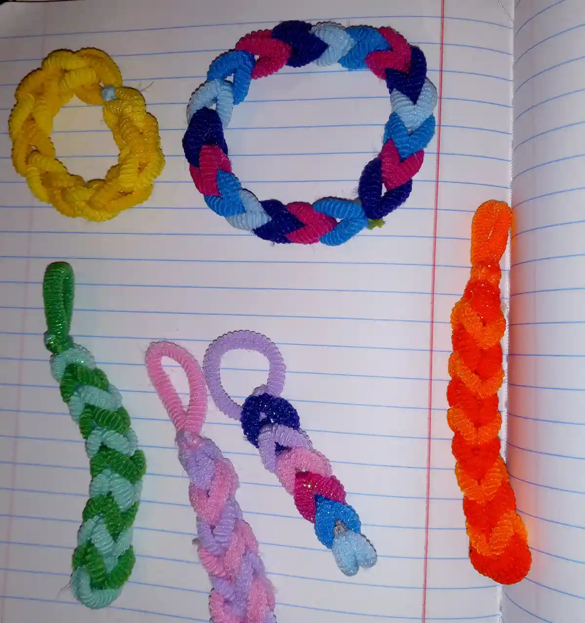 Five colorful, handcrafted braided items made from chenille stems are arranged on lined notebook paper to spell out the…