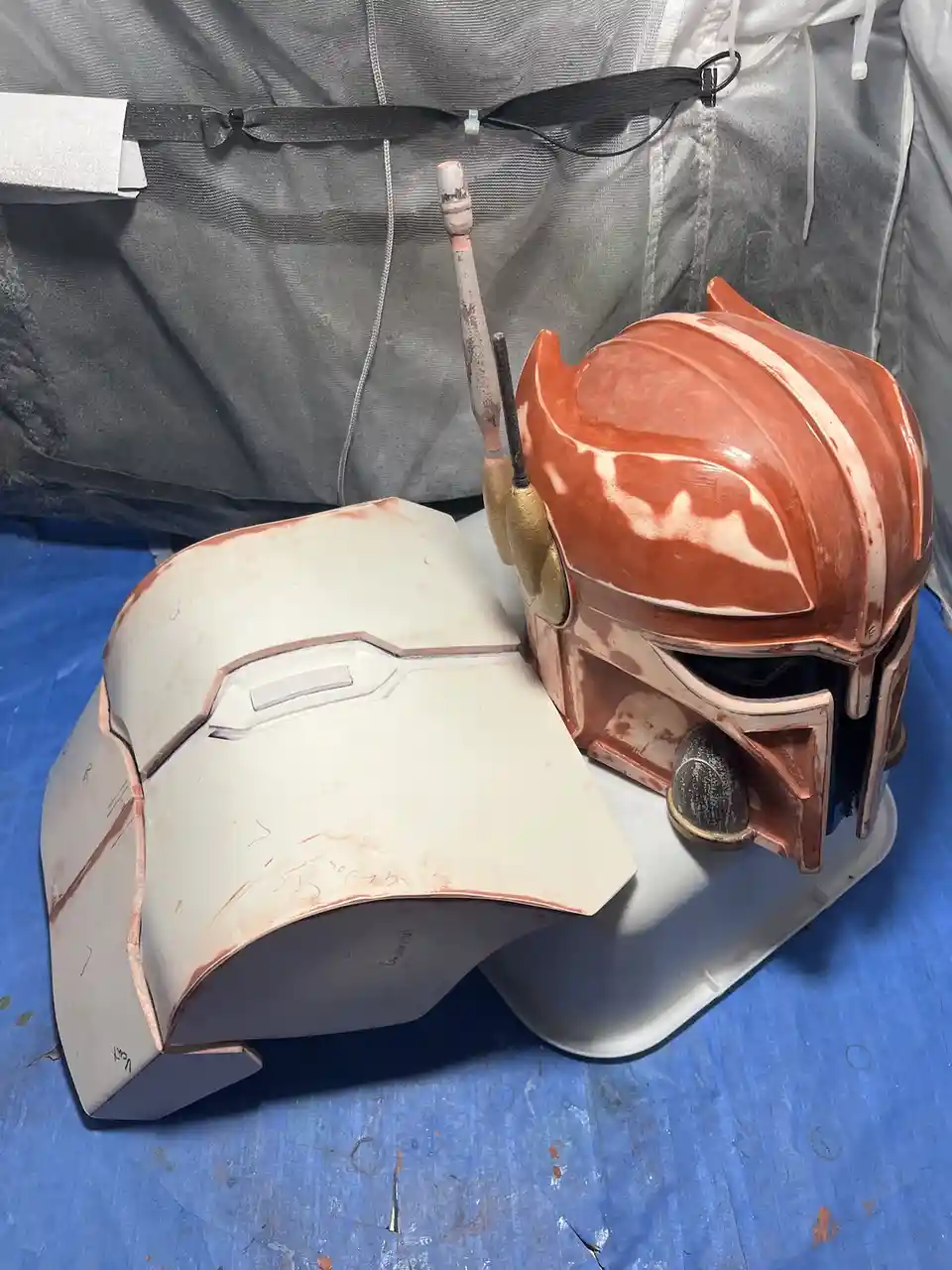Two partially painted Star Wars Mandalorian helmets sit on a white tray on a blue work surface. One helmet is mostly white…