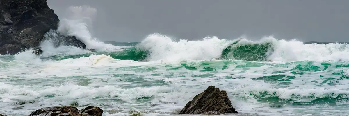 A dramatic photograph of powerful ocean waves crashing against dark, jagged rocks under a grey sky. The water is turbulent…