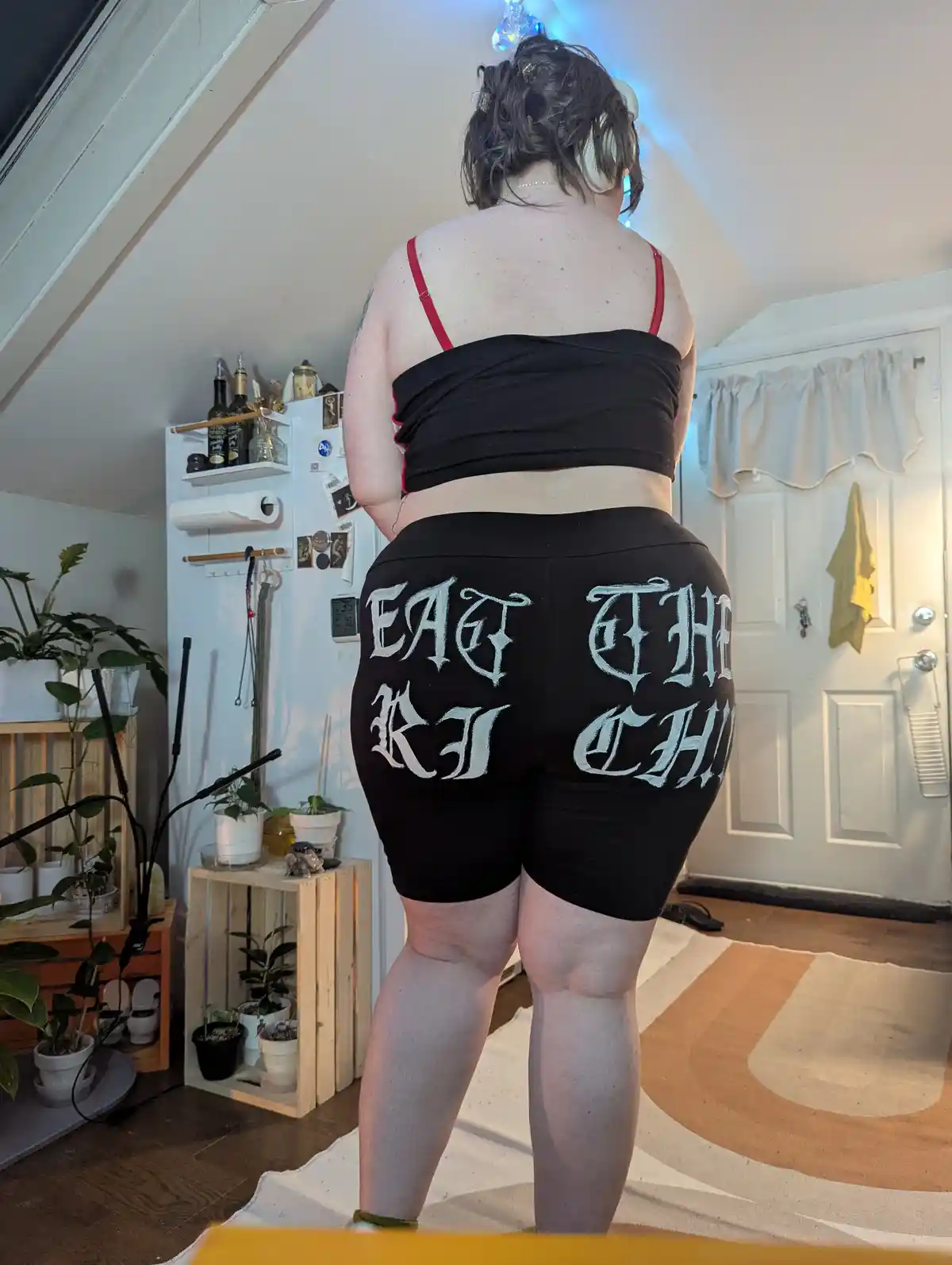 A person stands with their back to the camera in a room, wearing black shorts with the phrase 'EAT THE RICH' printed in…