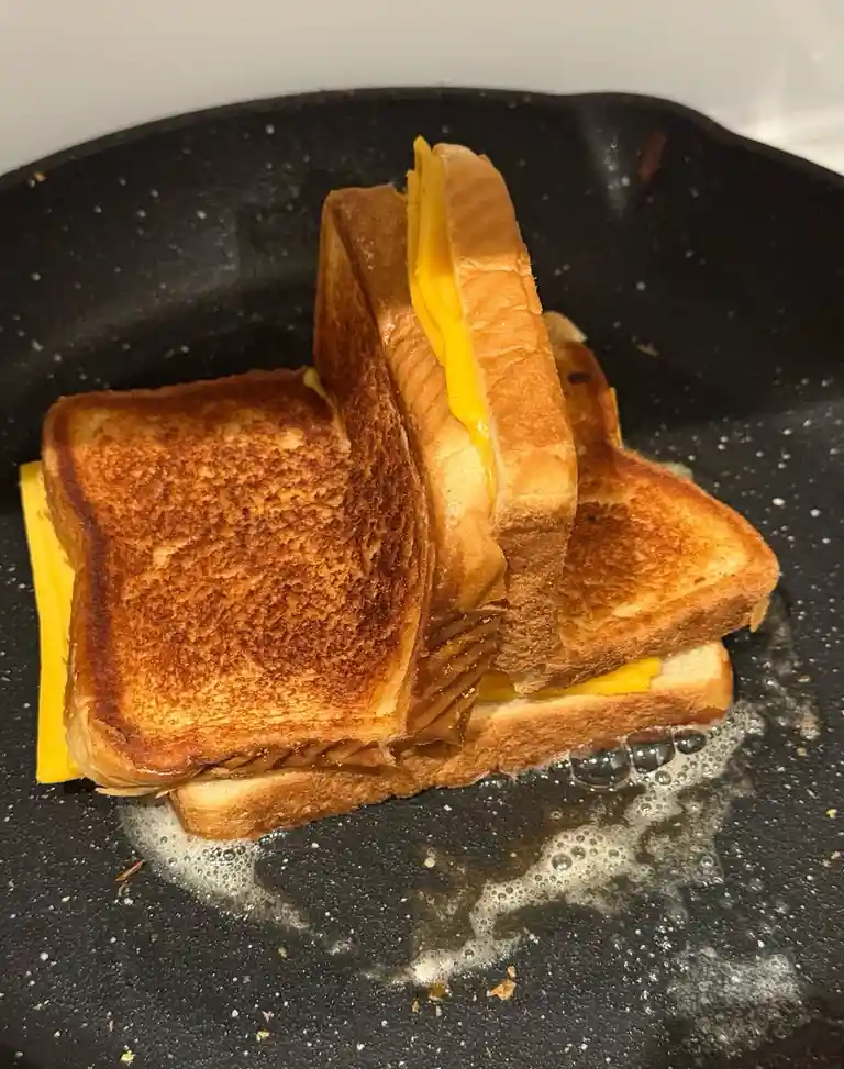 A grilled cheese sandwich is shown in a black frying pan, with the bread toasted golden brown and melted yellow cheese…