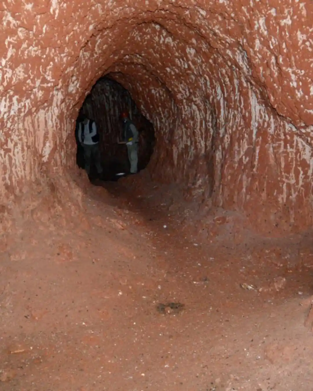 Two people stand inside a narrow, reddish-brown tunnel with rough, textured walls. One person wears a red hard hat and…