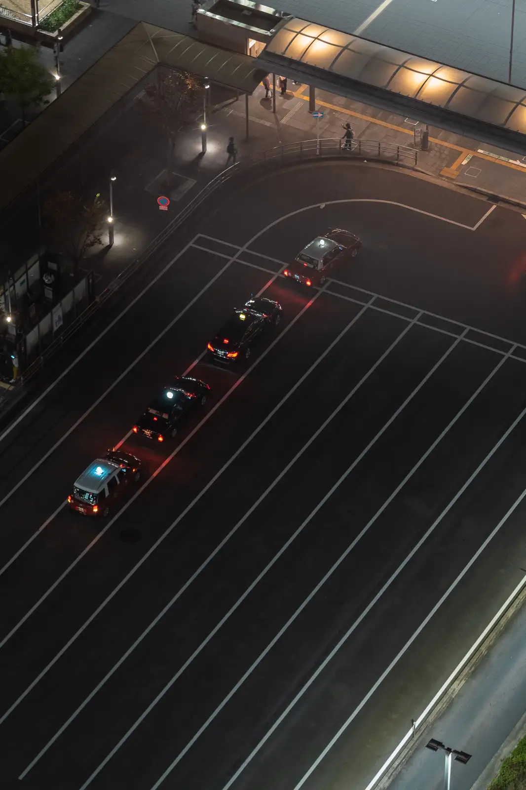 An aerial night view shows a line of taxis with illuminated roof lights parked in a designated taxi stand, with a few…