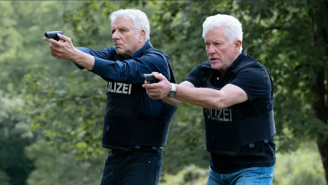 Two older men with white hair, both wearing vests labeled 'POLIZEI', are aiming handguns forward in a wooded area. The man…