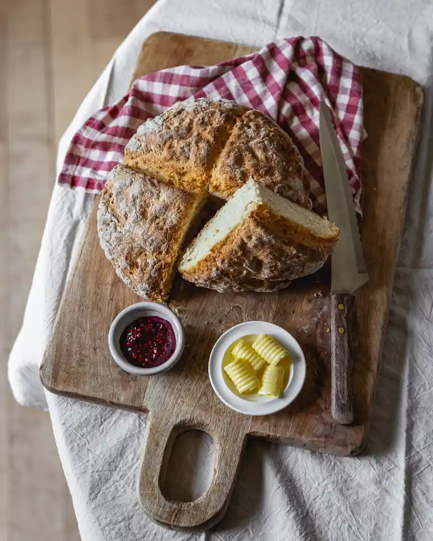 A rustic round loaf of Irish soda bread sits sliced on a wooden cutting board, accompanied by a knife, a red-and-white…