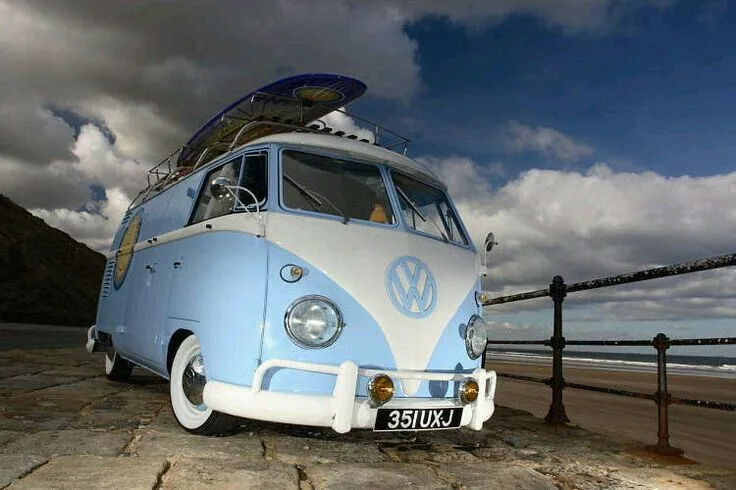 A light blue and white vintage Volkswagen Type 2 camper van is parked on a stone pier overlooking a beach with a surfboard…
