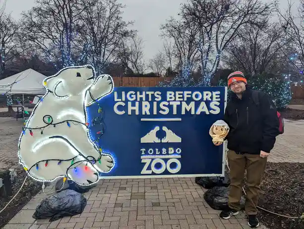 A man in a winter hat and jacket stands beside a large blue sign that reads 'LIGHTS BEFORE CHRISTMAS' and 'TOLEDO ZOO', next…