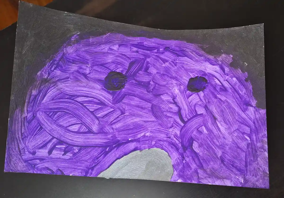 A child's painting on black paper depicts a purple, textured monster face with two black circular eyes and a gray…