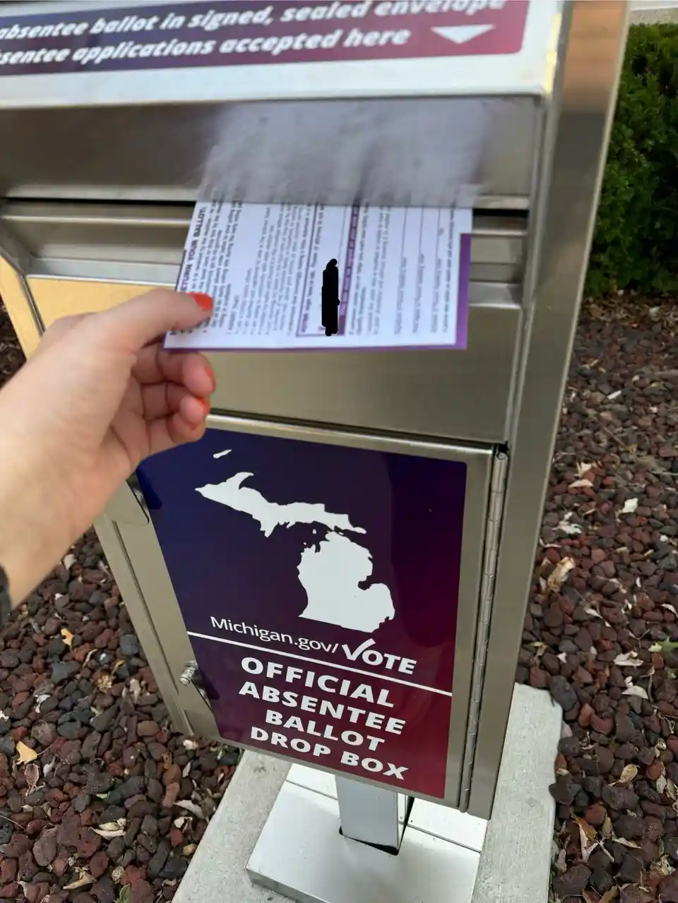 A person's hand with orange nail polish holds a paper ballot over a Michigan official absentee ballot drop box. The ballot…
