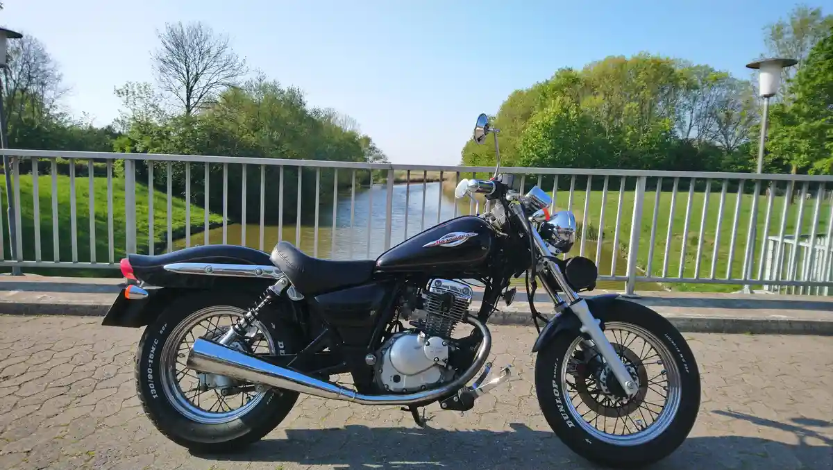 A black Suzuki motorcycle with chrome accents is parked on a paved path beside a metal railing overlooking a calm river and…