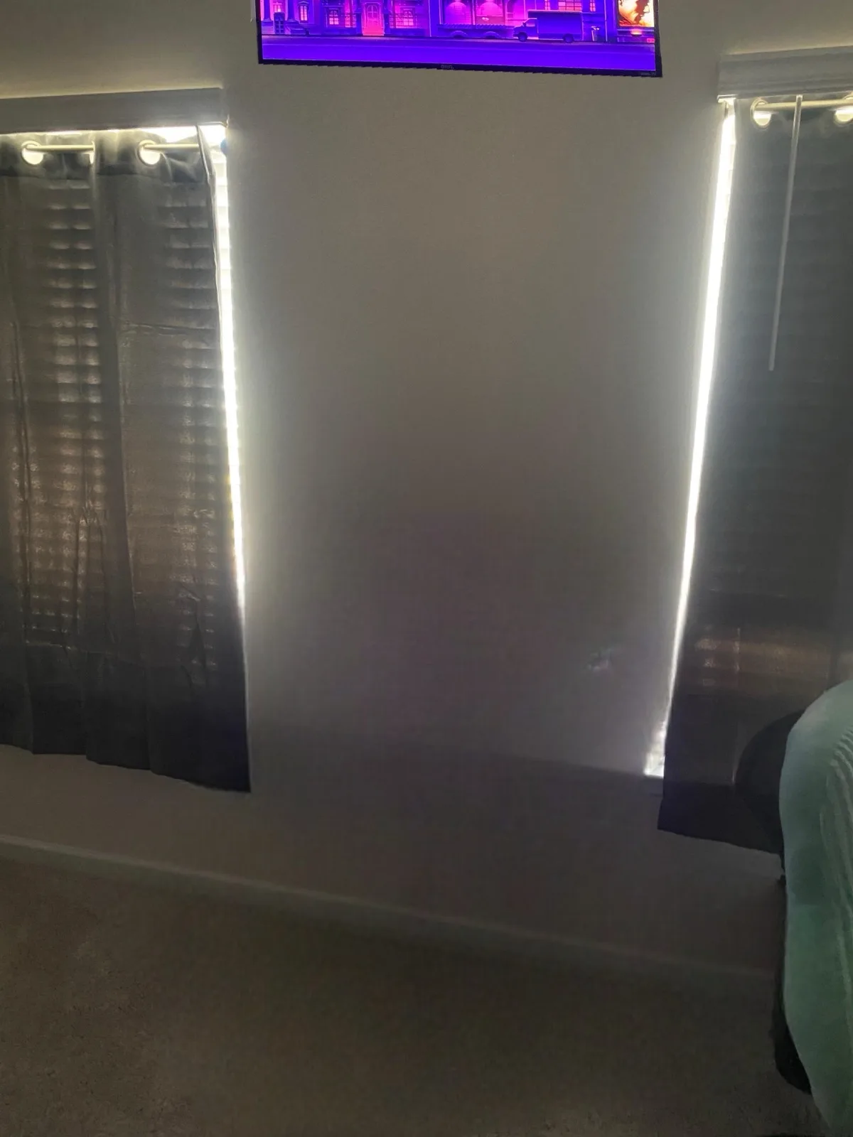 A dimly lit bedroom with a TV mounted on the wall displaying a purple-hued scene, flanked by two vertical strips of bright…