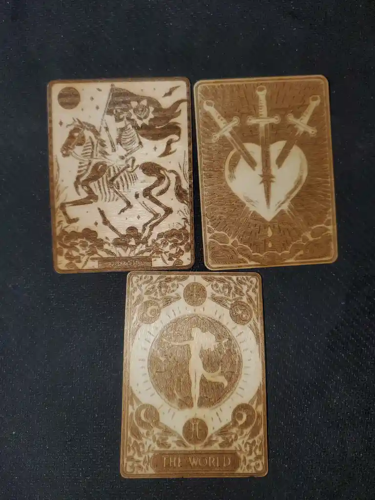 Three wooden tarot cards are laid out on a dark surface: 'Death' showing a skeletal figure on a horse, 'The World' depicting…