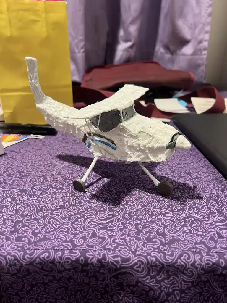 A handmade, crudely crafted model airplane made of white textured material sits on a purple patterned tablecloth. The plane…